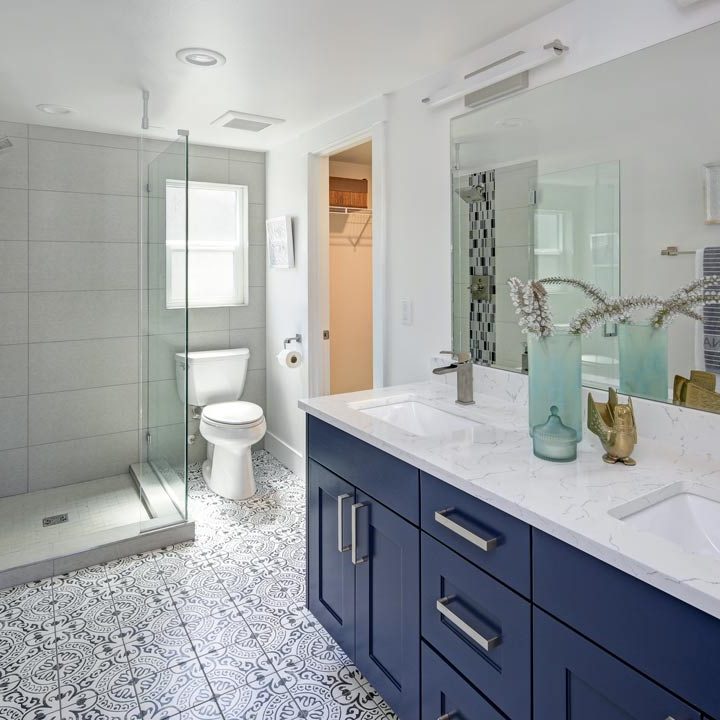 A modern bathroom with blue cabinets, patterned tile flooring, marble countertop, a flush toilet, glass enclosed shower area, and a wide mirror